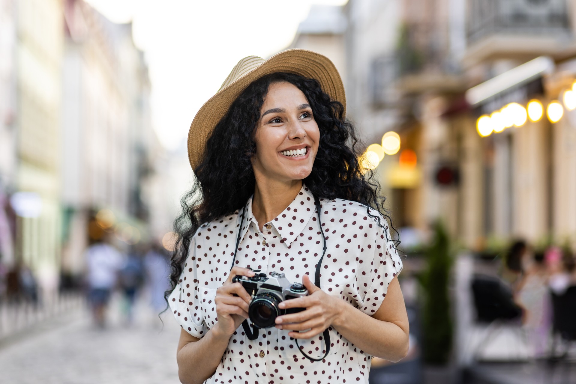 Young beautiful hispanic woman with curly hair walking in the evening city with a camera, female tourist on a trip exploring historical landmarks in the city